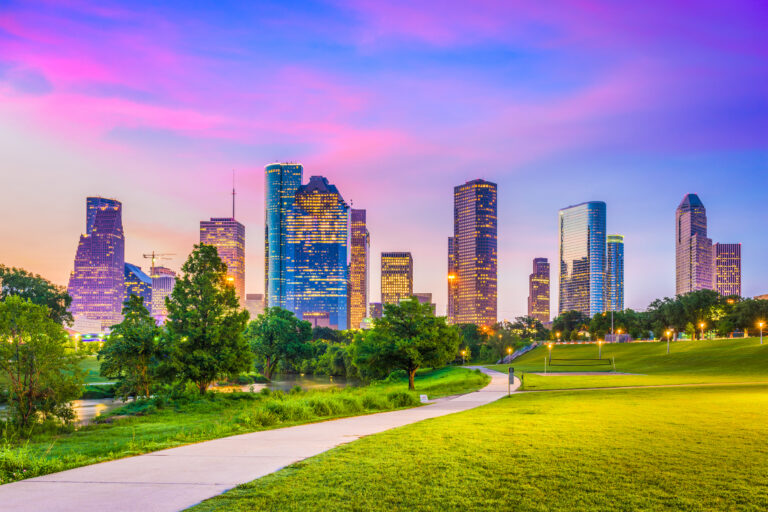 A vibrant twilight sky blankets the Houston, Texas, USA downtown city skyline, with illuminated buildings reflecting off the green spaces in the foreground.