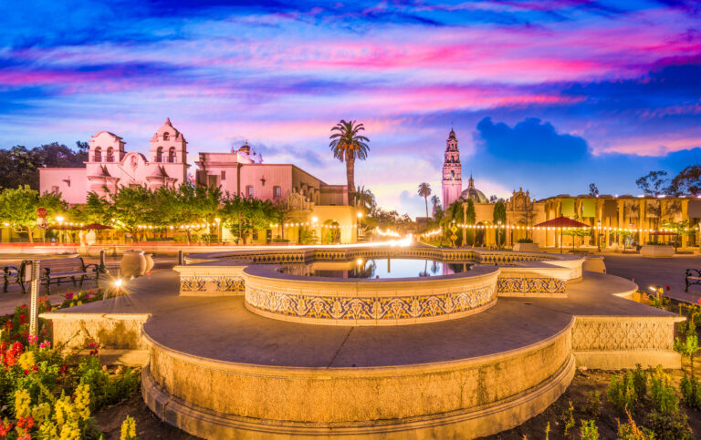 A vibrant sunset sky blankets a serene park and square in San Diego, California, highlighting the area’s picturesque architecture and tranquil fountain.