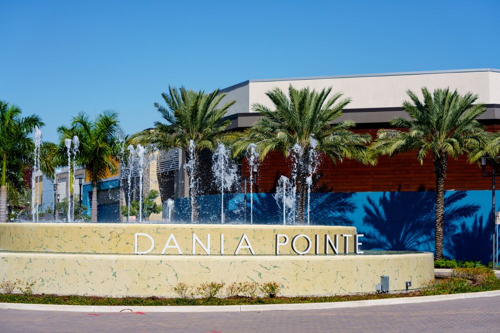 Dania pointe written in white letters with water fountains and palm trees behind.