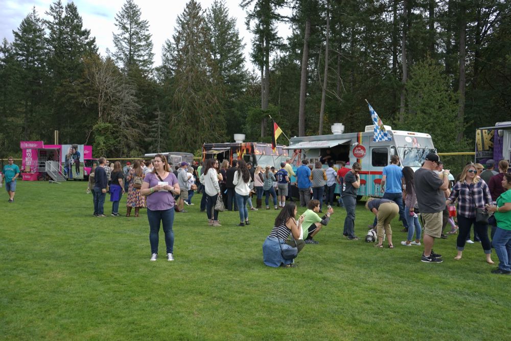 A flock of people standing near food trucks during a cider festival at the park.