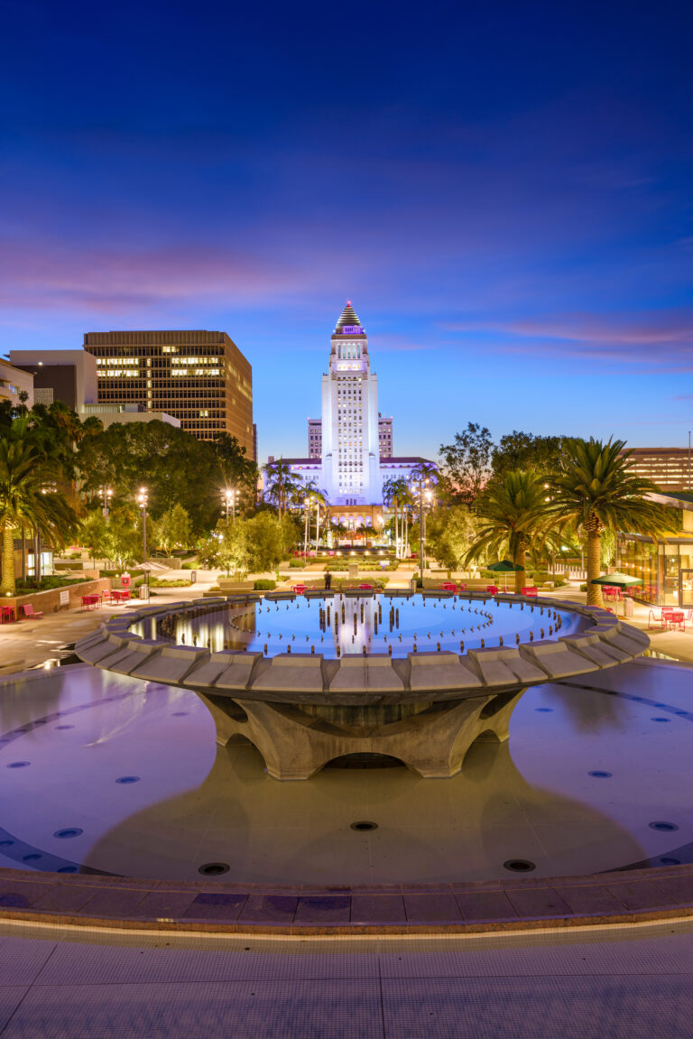 A serene twilight view of the Los Angeles City Hall, framed by the vibrant evening sky and the reflective waters of the foreground fountain.