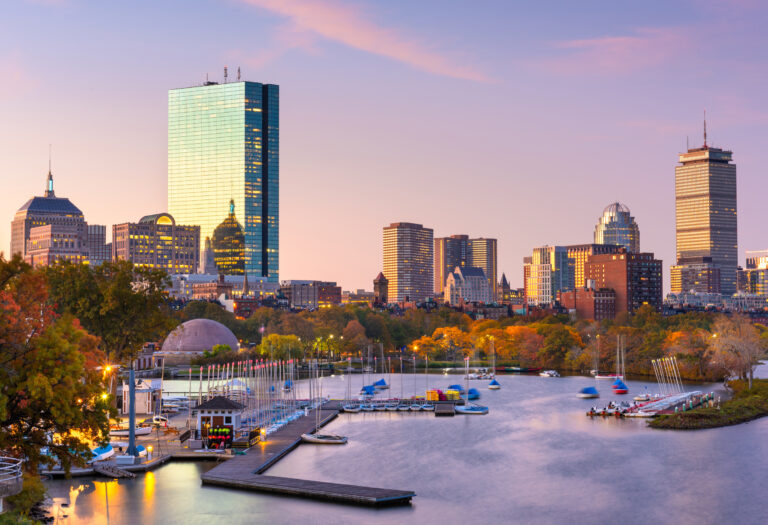 A serene dawn breaks over the Boston skyline, reflecting off the Charles River and highlighting the city’s architecture and moored sailboats.