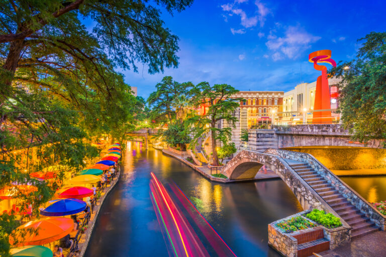 A vibrant evening view of the San Antonio, Texas, USA cityscape along the bustling River Walk, highlighted by colorful umbrellas and streaks of light from a passing boat.