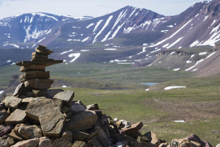 A serene cairn stands as a guide on the verdant slopes leading to King’s Peak, Utah’s highest summit, against a backdrop of snow-streaked mountains.