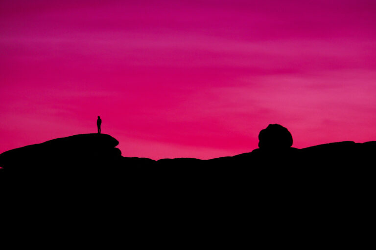 A solitary figure stands atop a rocky outcrop against the vibrant hues of a sunset sky in Joshua Tree National Park, California, USA.