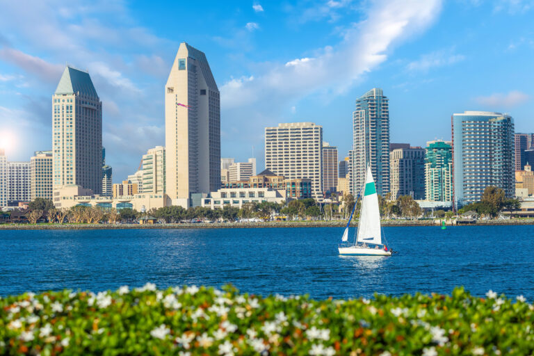 A sailboat cruises by the vibrant Downtown San Diego skyline, showcasing the city’s dynamic architecture against a clear blue sky.
