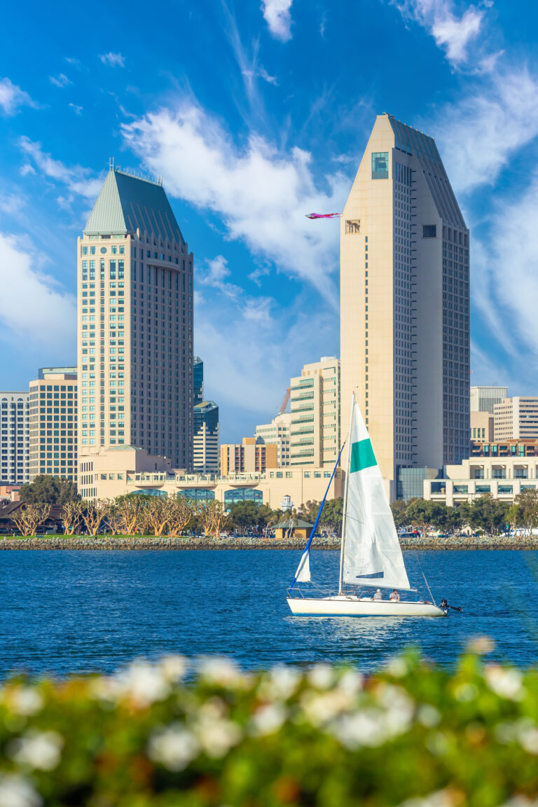 A sailboat glides across the water with the iconic towers of Downtown San Diego rising majestically in the background under a clear blue sky.