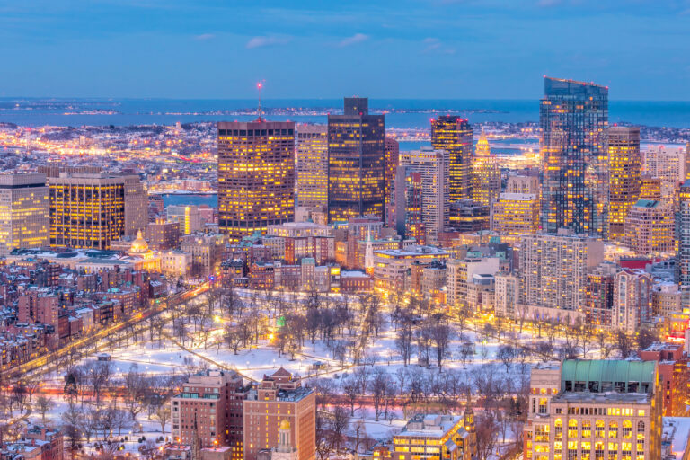 The Boston skyline glows warmly against the cool hues of winter sunset, with the snow-covered expanse of Boston Common park offering a serene foreground.