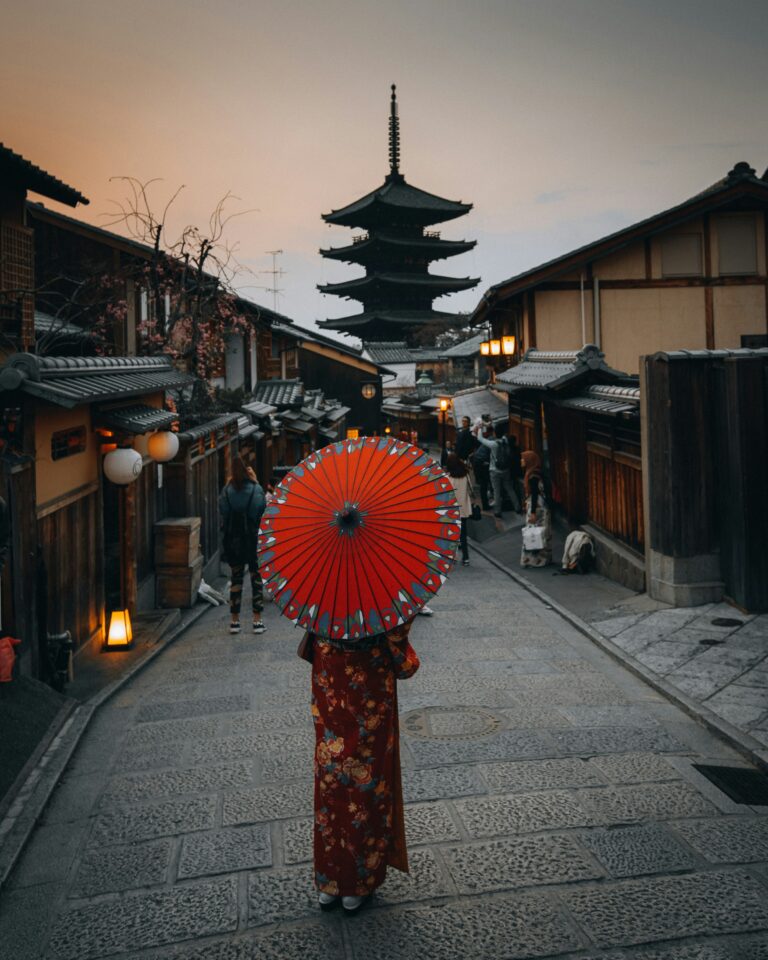 In the historic streets of Kyoto, a person in traditional kimono and holding a red umbrella creates a picturesque scene against the backdrop of the ancient five-tiered pagoda at dusk.