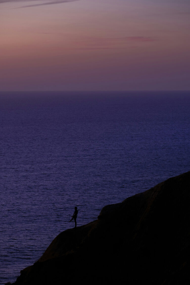 A solitary figure stands on a cliff overlooking the vast, tranquil ocean as the sky fades into twilight hues.