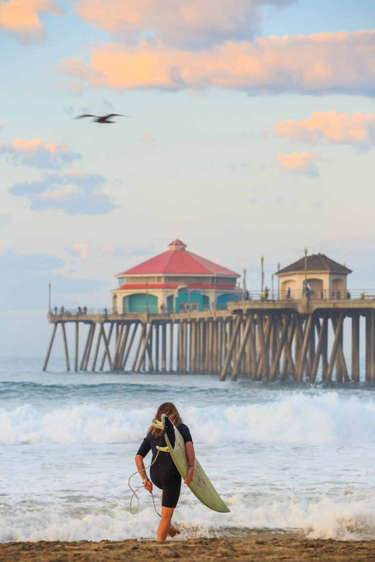 A surfer prepares to enter the waves at Huntington Beach with the iconic pier in the background, bathed in the soft glow of sunrise.
