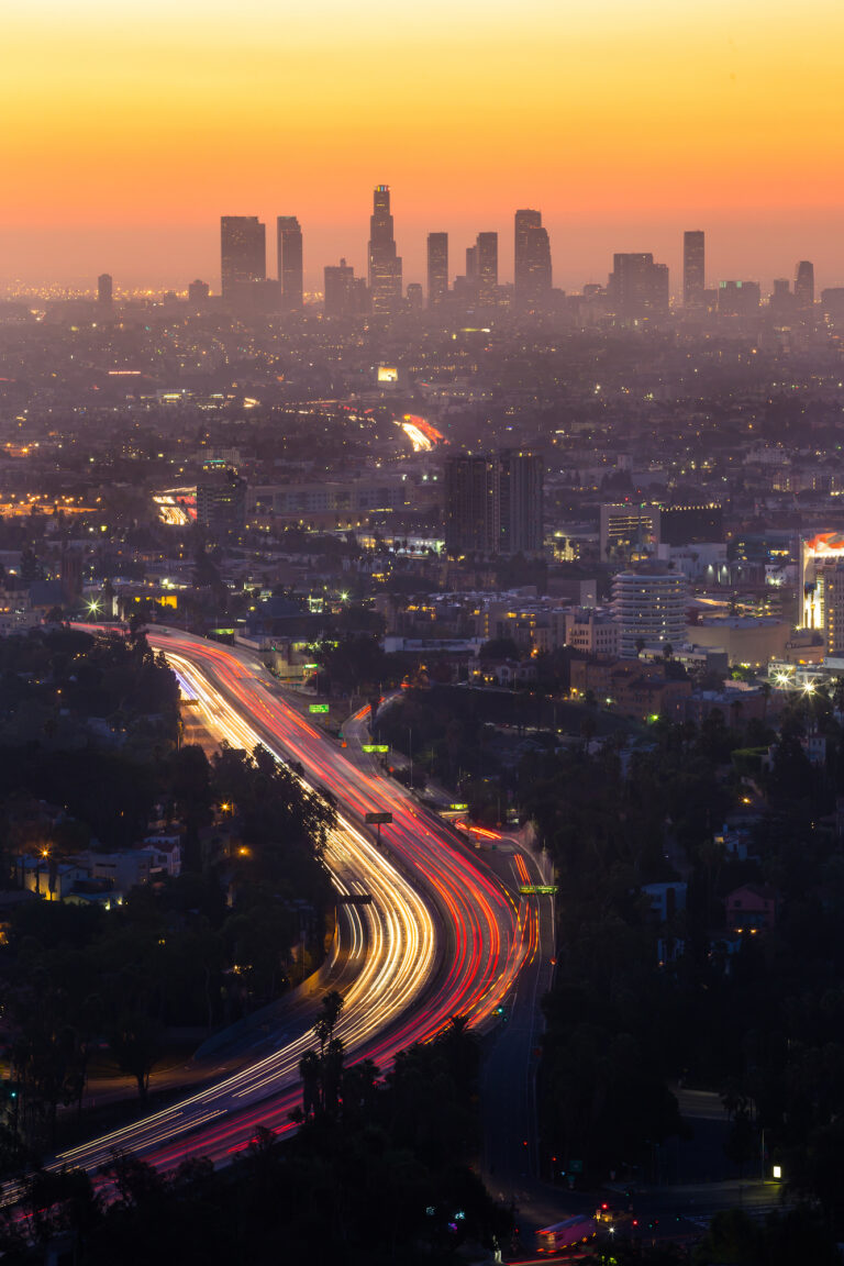 The vibrant hues of sunset cast a warm glow over the bustling Downtown Los Angeles skyline, with streaks of light tracing the city’s ever-moving traffic.